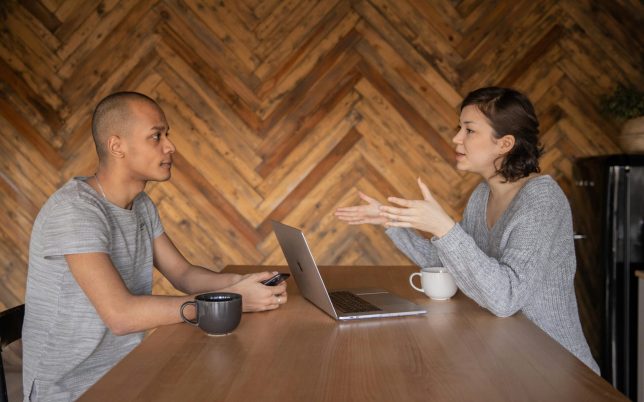 Focused woman explaining opinion to ethnic male coworker during business teamwork sitting at table with laptop and coffee cups in cozy kitchen against wooden wall and looking at each other